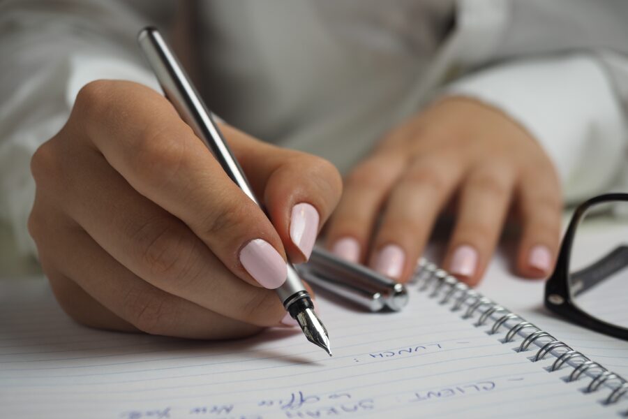 Woman in White Long Sleeved Shirt Holding a Pen Writing on a Paper