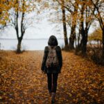 woman walking on pathway with falling leaves near body of water during daytime